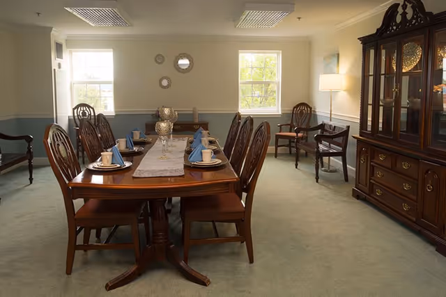 A formal dining room with a long wooden dining table set for eight people with blue napkins, cups, and plates. The room has two windows letting in natural light, a wooden china cabinet on the right, and several wooden chairs around the table and against the walls. The walls are painted light beige with a blue-gray wainscoting, and there is a floor lamp in the corner.