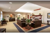 A spacious and well-lit living room area in a senior living facility featuring comfortable brown sofas and armchairs arranged around a central coffee table with a plant. The room has a fireplace with a mantel decorated with plants and artwork on the walls. There is a reception desk visible in the background, along with additional seating and decorative elements.
