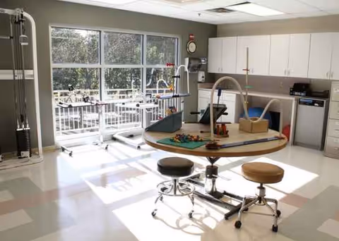 Rehabilitation therapy room with round table, stools, play equipment, large windows, and cabinets.