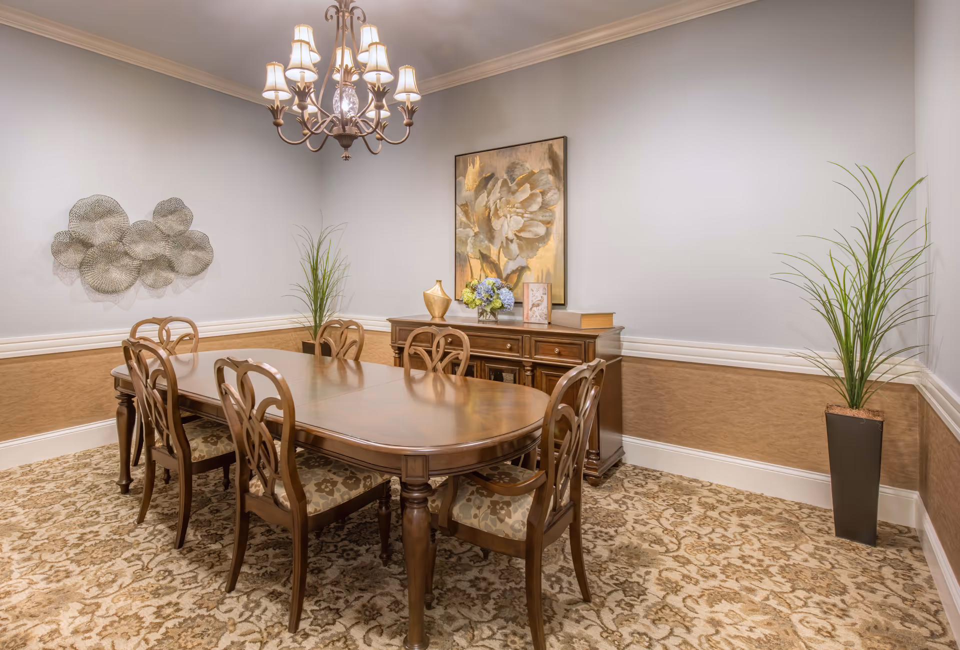 A formal dining room with a wooden dining table and six matching chairs with patterned cushions. The room features a chandelier hanging above the table, a sideboard with decorative items including a vase with flowers, a framed picture, and a book. The walls are painted light gray with a beige wainscoting, and there are two tall potted plants in the corners. The floor is covered with a patterned carpet.