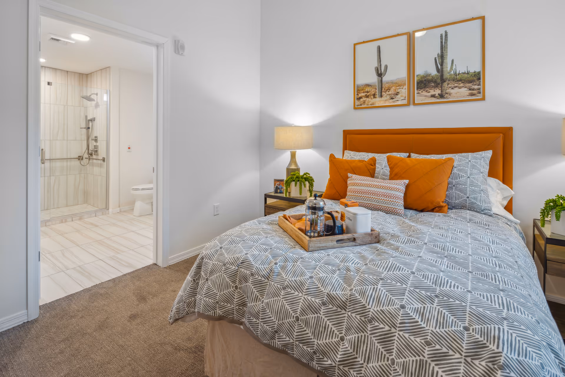 A cozy bedroom with a patterned gray and white bedspread, orange accent pillows, and a matching orange headboard. There is a wooden tray on the bed with a French press, a cup, and some snacks. Two framed pictures of cacti hang above the bed. On either side of the bed are nightstands with lamps and small green plants. An open door reveals a bathroom with a walk-in shower and a toilet.