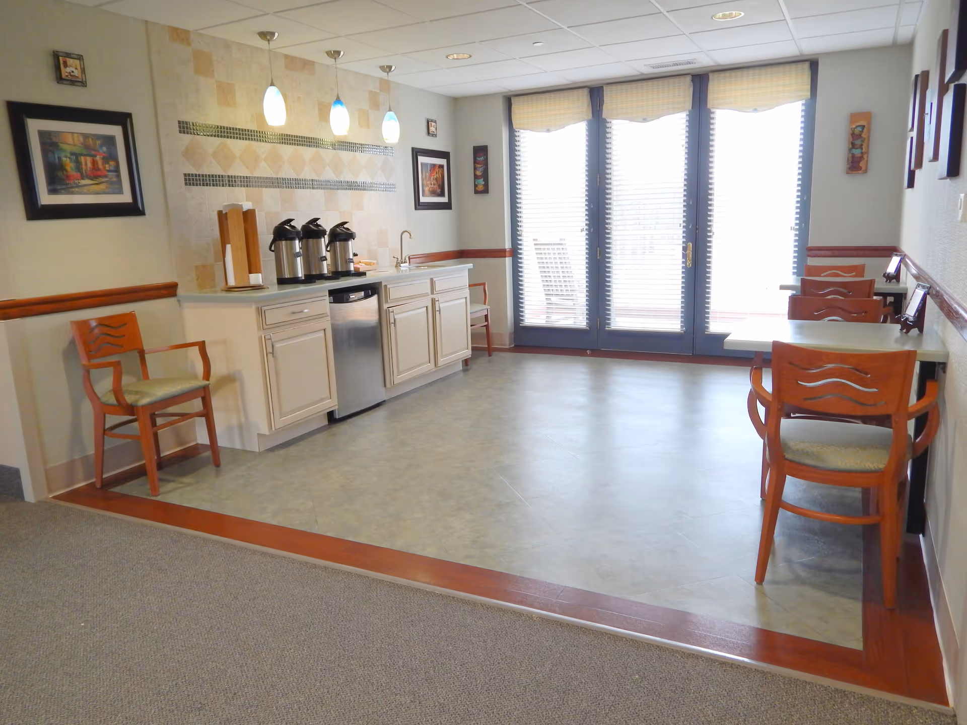 A small dining area with a kitchenette featuring a countertop, sink, and three coffee dispensers. There are wooden chairs with cushions placed around small tables and along the walls. The room has large glass doors with blinds letting in natural light, and framed artwork on the walls.