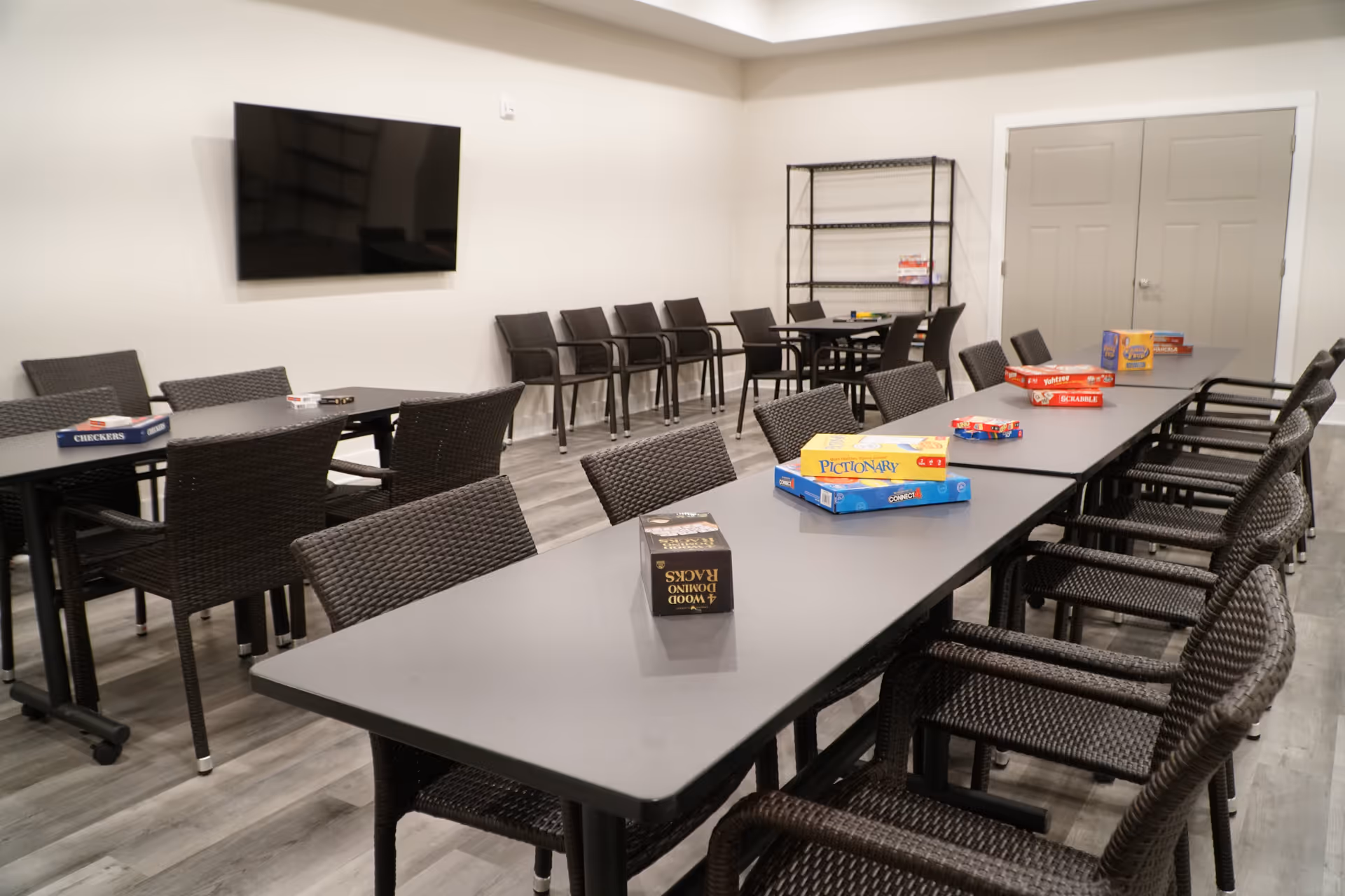 A game room with multiple black tables and black woven chairs arranged around them. Various board games such as Pictionary, Scrabble, Checkers, and Dominoes are placed on the tables. A large flat-screen TV is mounted on the wall, and there is a black metal shelving unit in the corner with more games. The room has light-colored walls and wood-patterned flooring.