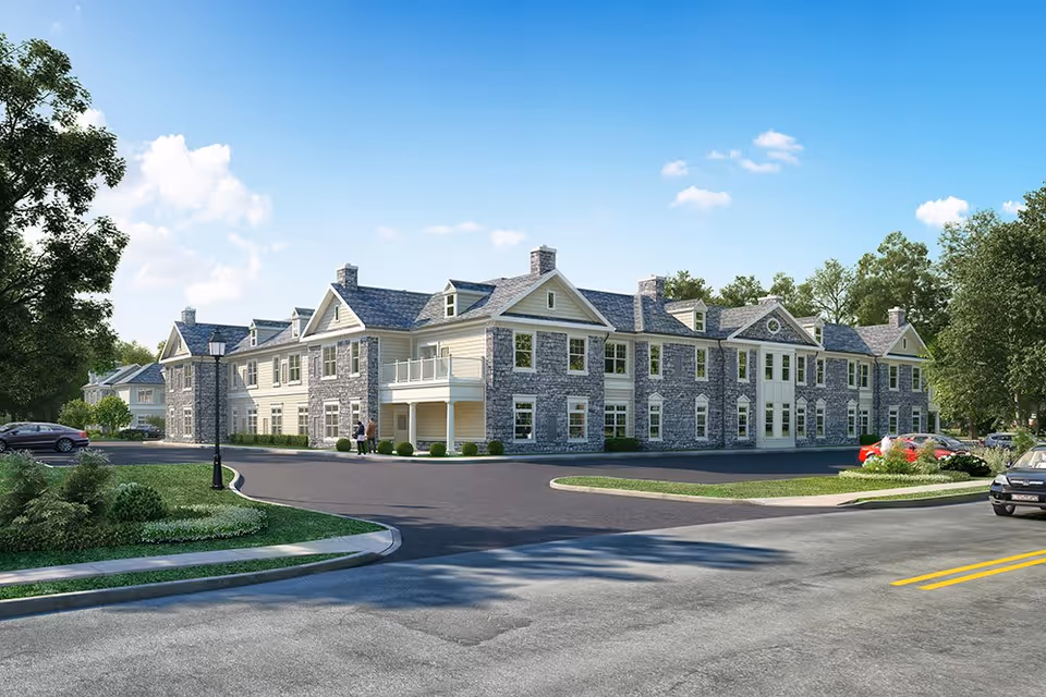 Two-story stone-and-siding senior living building with a parking lot, landscaped grounds, and cars under a clear blue sky.