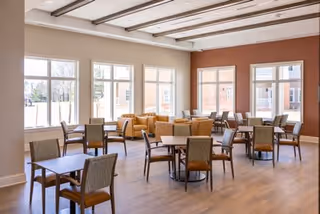 Bright dining room with multiple tables and chairs and lounge seating by large windows.