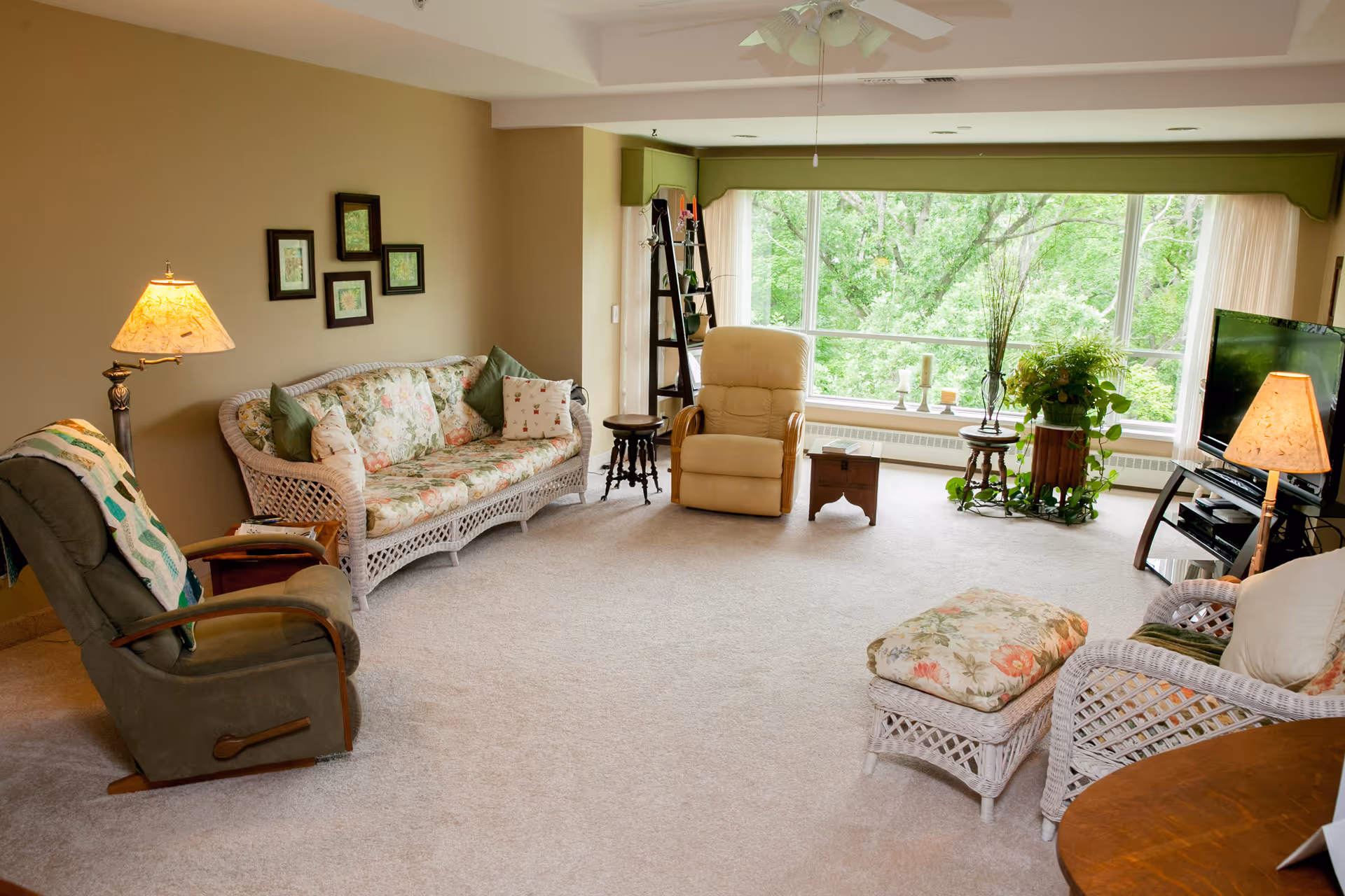 A cozy living room with a large window showing green trees outside. The room features a floral-patterned wicker sofa and matching ottoman, a beige recliner, a green armchair with a quilt draped over it, a wooden side table with a lamp, a TV on a stand with another lamp, and various plants and decorative items near the window.