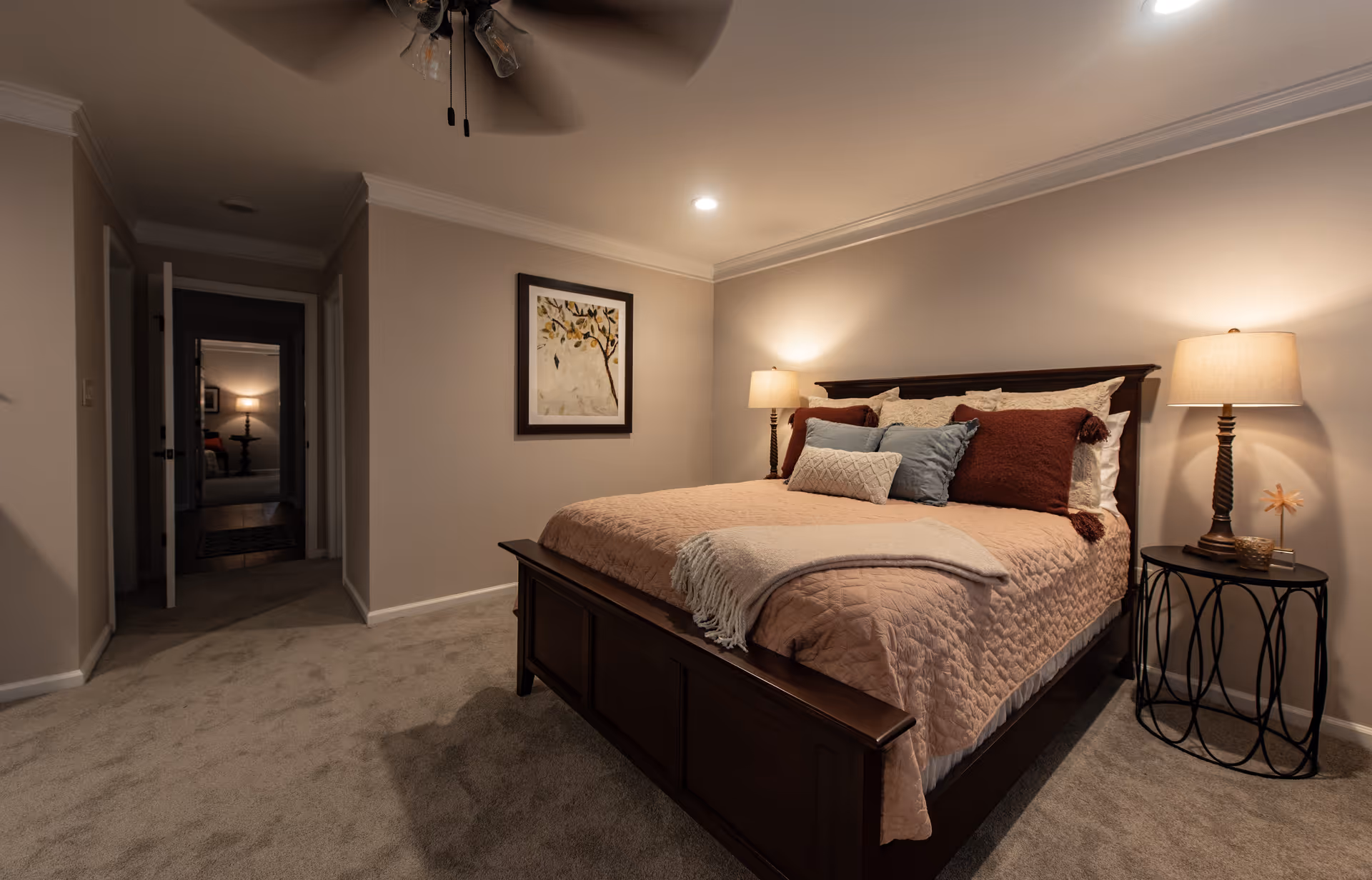 A cozy bedroom with a large wooden bed featuring a beige quilt, multiple pillows in shades of brown and blue, and a beige throw blanket. Two bedside tables with matching lamps provide warm lighting. A framed artwork hangs on the wall above the bed. The room has beige carpet flooring and neutral-colored walls. An open doorway leads to another room with a visible lamp and furniture.