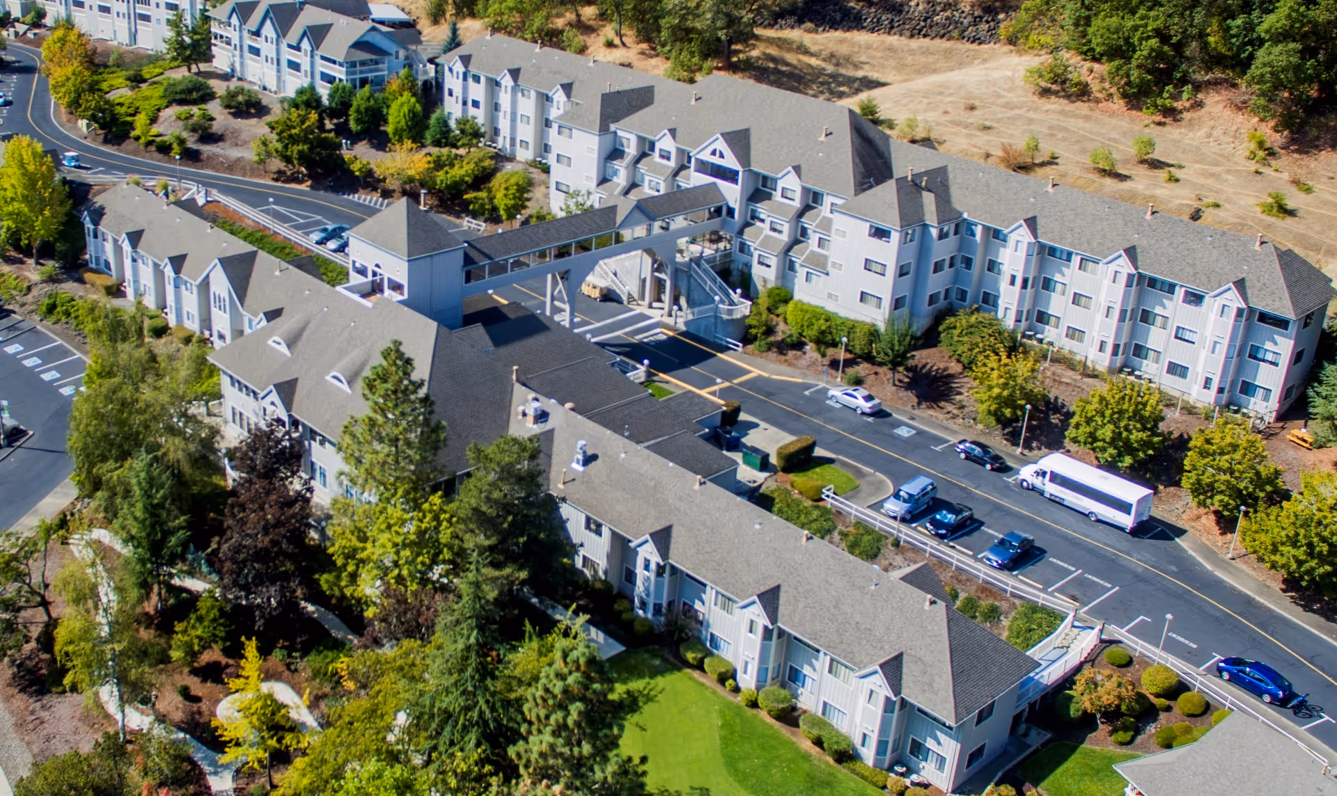Aerial view of Linus Oakes Village senior living facility showing multiple connected buildings with gray roofs, a covered walkway bridge, surrounding parking lots with cars, and landscaped green areas with trees and shrubs.
