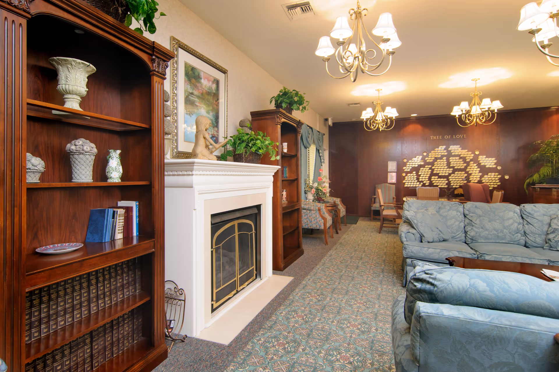 A cozy living room area in a senior living facility featuring a white fireplace with decorative items on the mantel, wooden bookshelves filled with books and ornaments, blue upholstered sofas, patterned carpet, and elegant chandeliers hanging from the ceiling. A wall in the background displays a 'Tree of Love' plaque with names, and there are several chairs and plants adding to the warm ambiance.