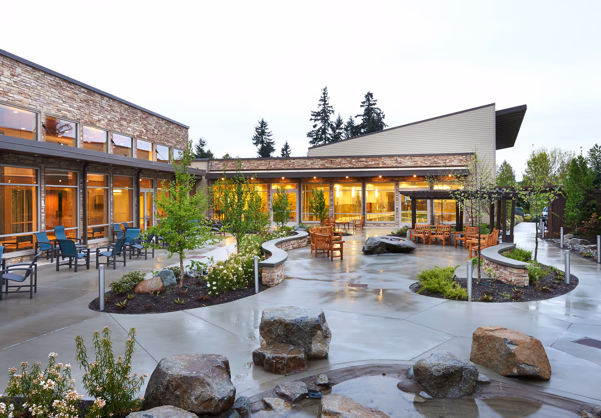 Outdoor courtyard area of a senior living facility with wet concrete pathways, landscaped garden beds, large rocks, small trees, and multiple seating areas with wooden benches and chairs. The building has large windows and stone and siding exterior walls, with warm interior lighting visible through the windows.