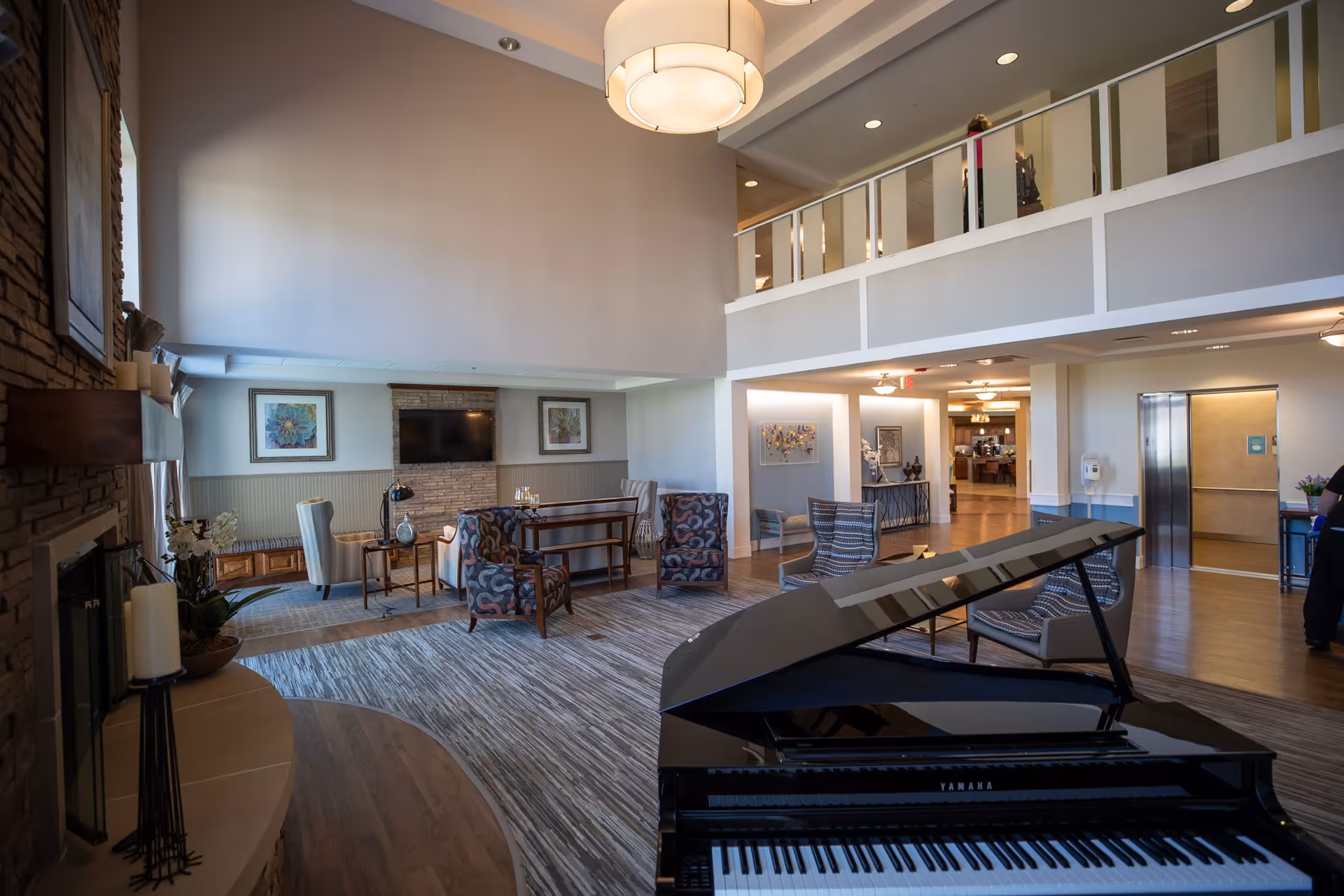 A spacious and well-lit common area in a senior living facility featuring a black Yamaha grand piano in the foreground, several patterned armchairs arranged around a carpeted area, a fireplace with candles on the left, and a wall-mounted TV above a stone accent wall. The room has a high ceiling with a large hanging light fixture and a second-floor balcony overlooking the space. In the background, there is an open hallway leading to other parts of the facility.