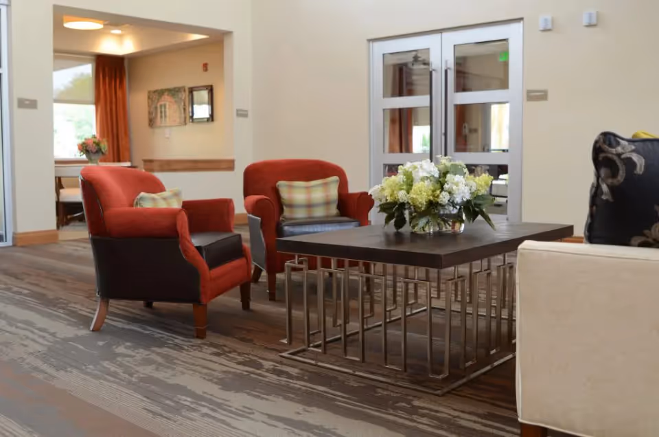 A cozy seating area in a senior living facility with two red armchairs, a beige sofa, and a dark wooden coffee table with a floral arrangement. The room has neutral-colored walls, a patterned carpet, and double glass doors in the background.