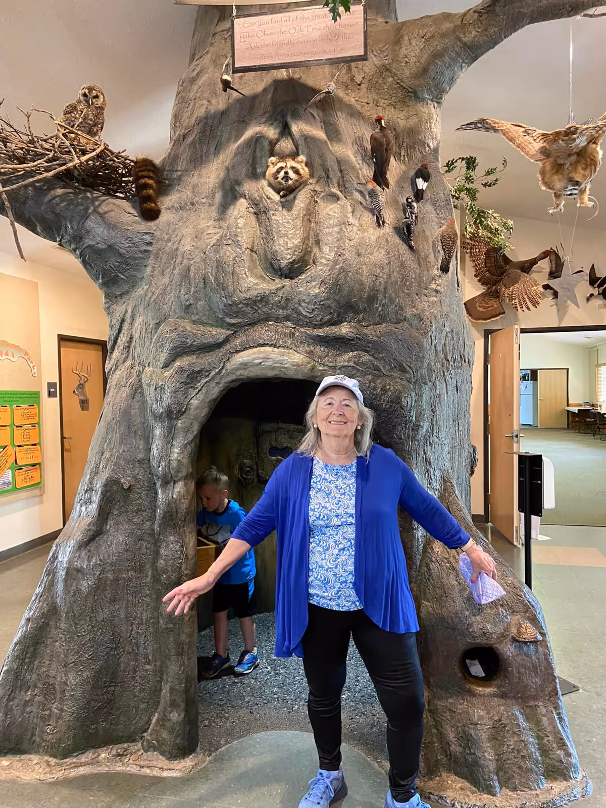 An elderly woman wearing a blue cardigan, patterned shirt, black pants, and a white cap stands smiling with arms outstretched in front of a large artificial tree structure. The tree has various animal figures attached, including a raccoon peeking out of a hole, birds, and an owl. A young boy is partially visible inside the hollow base of the tree. The setting appears to be an indoor facility with open doors and informational displays on the walls.