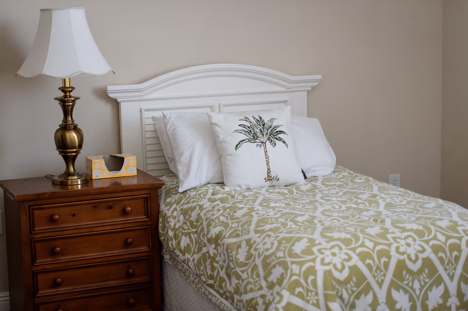 A neatly made single bed with a white headboard, white pillows, and a decorative pillow featuring a palm tree design. The bedspread has a green and white floral pattern. Next to the bed is a wooden nightstand with three drawers, a brass table lamp with a white lampshade, and a yellow tissue box.