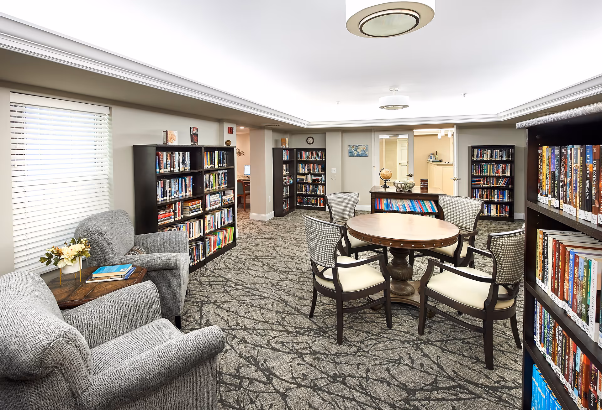 Well-lit senior living library/lounge with bookshelves, a round table surrounded by chairs, and upholstered armchairs by a window.