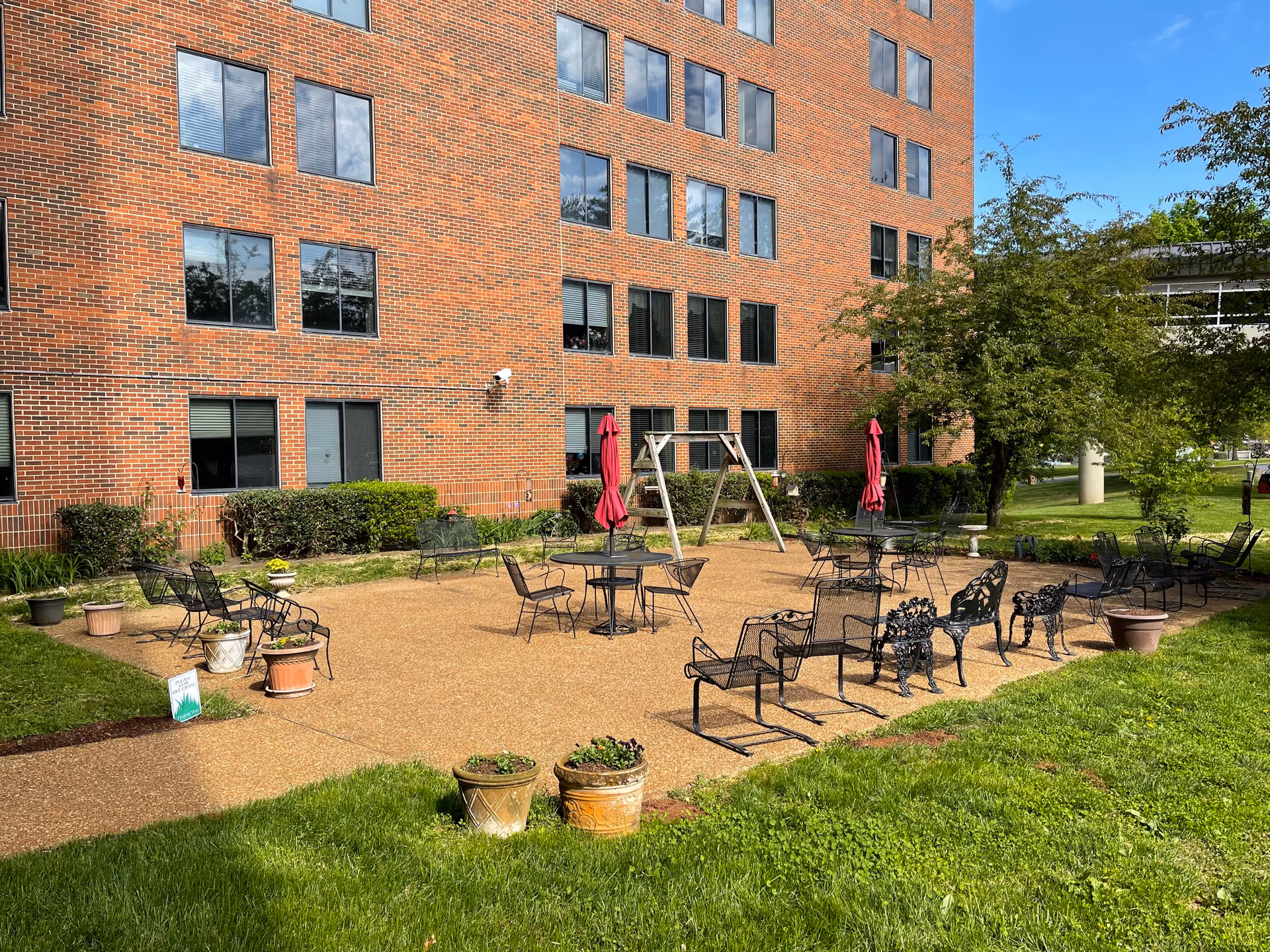 Outdoor patio area at The McKendree retirement home featuring multiple metal chairs and tables with closed red umbrellas, surrounded by potted plants and green grass, adjacent to a multi-story brick building under a clear blue sky.