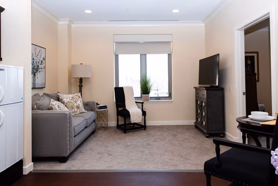 A cozy living room in a senior living facility featuring a gray sofa with patterned cushions, a black armchair with a white throw, a window with a potted plant on the sill, a dark wooden TV stand with a flat-screen TV, a floor lamp, and a small round side table. The room has beige walls and carpeted flooring.