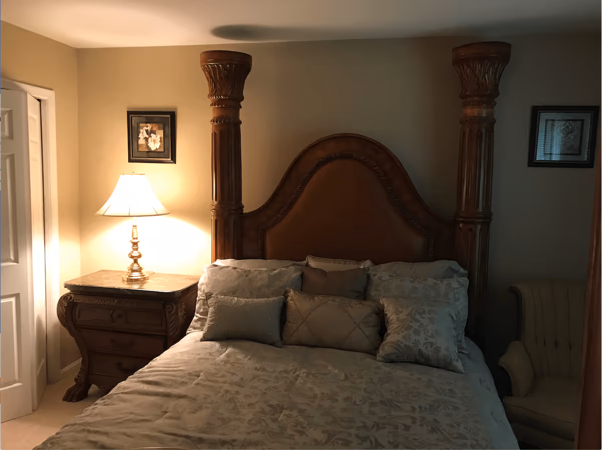 A bedroom with a large wooden bed featuring an ornate headboard and multiple decorative pillows. To the left of the bed is a wooden nightstand with a marble top and a lit table lamp. There are two framed pictures on the wall, one above the nightstand and one to the right of the bed. A beige upholstered armchair is partially visible on the right side of the image.