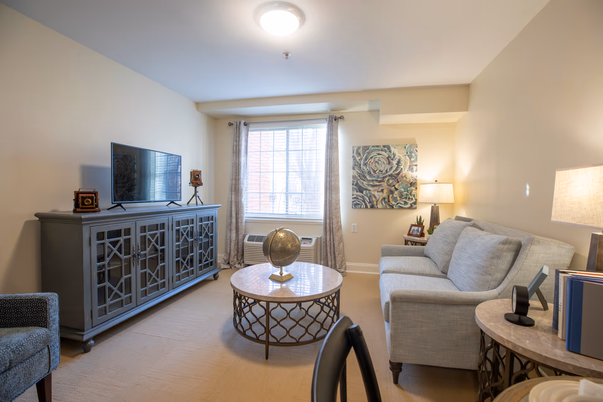 A cozy living room with a light gray sofa, a round coffee table with a decorative globe, a gray cabinet with a flat-screen TV on top, and a window with curtains letting in natural light. There are two table lamps and a floral wall art piece on the wall.