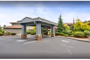 Covered entrance area of a retirement and assisted living facility with a paved driveway, landscaped greenery including bushes and trees, and a building with a brown roof in the background under a clear sky.