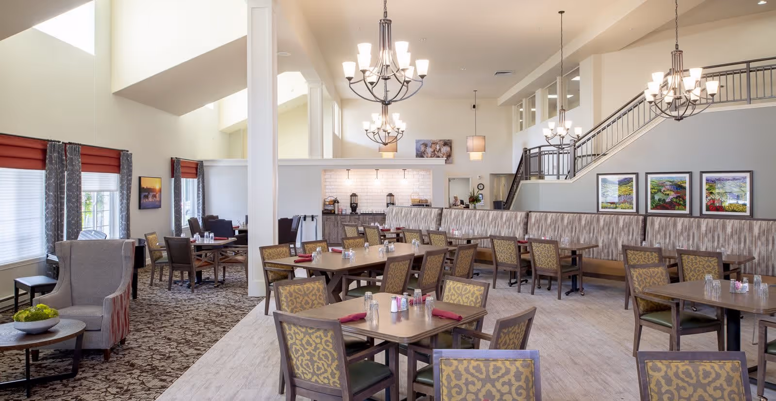 A spacious and well-lit dining area in a senior living facility with multiple tables and chairs arranged neatly. The room features high ceilings with modern chandeliers, large windows with red and gray curtains, patterned carpet and light wood flooring. There is a seating area with armchairs and a small table on the left, and a counter area with coffee machines and other amenities in the back. A staircase with a railing is visible on the right side, along with framed artwork on the walls.