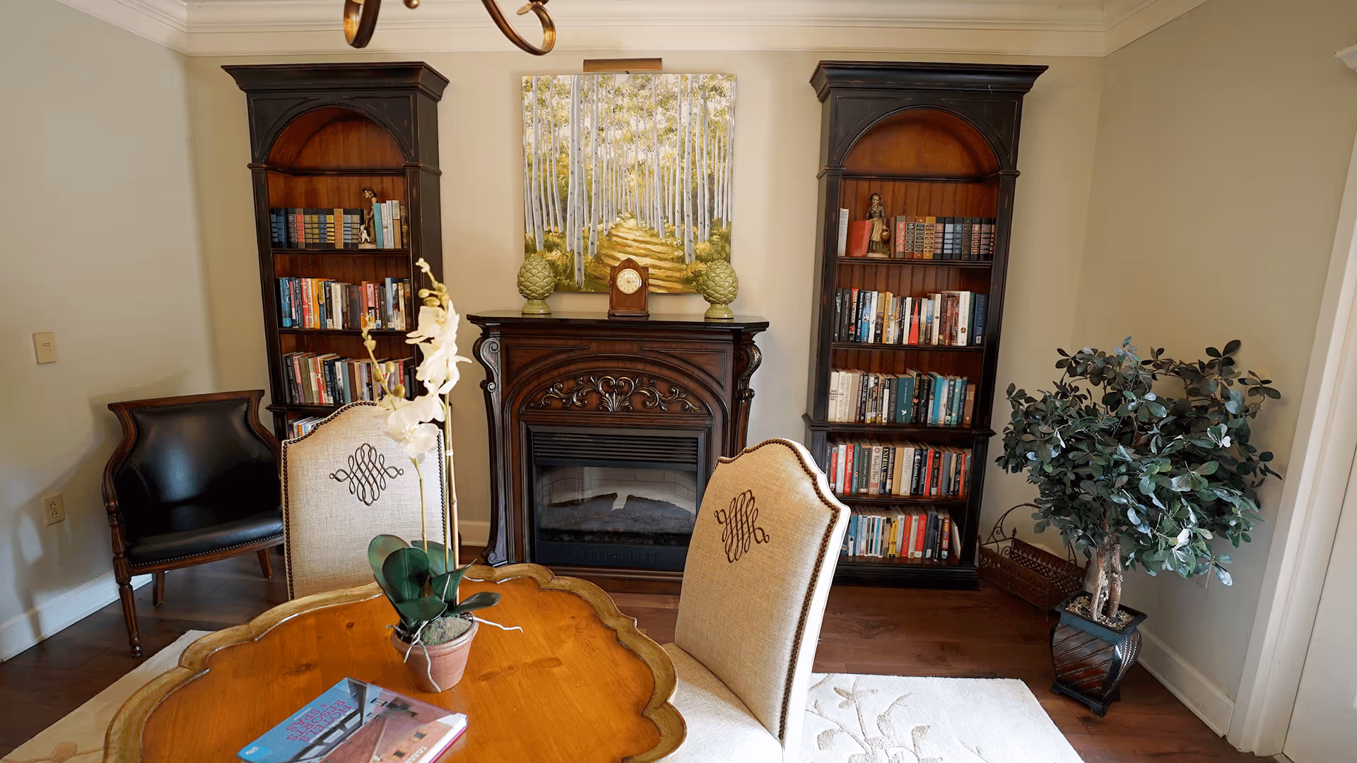 Cozy living room with a round wooden table and upholstered chairs facing a decorative fireplace flanked by two bookcases and a potted plant.