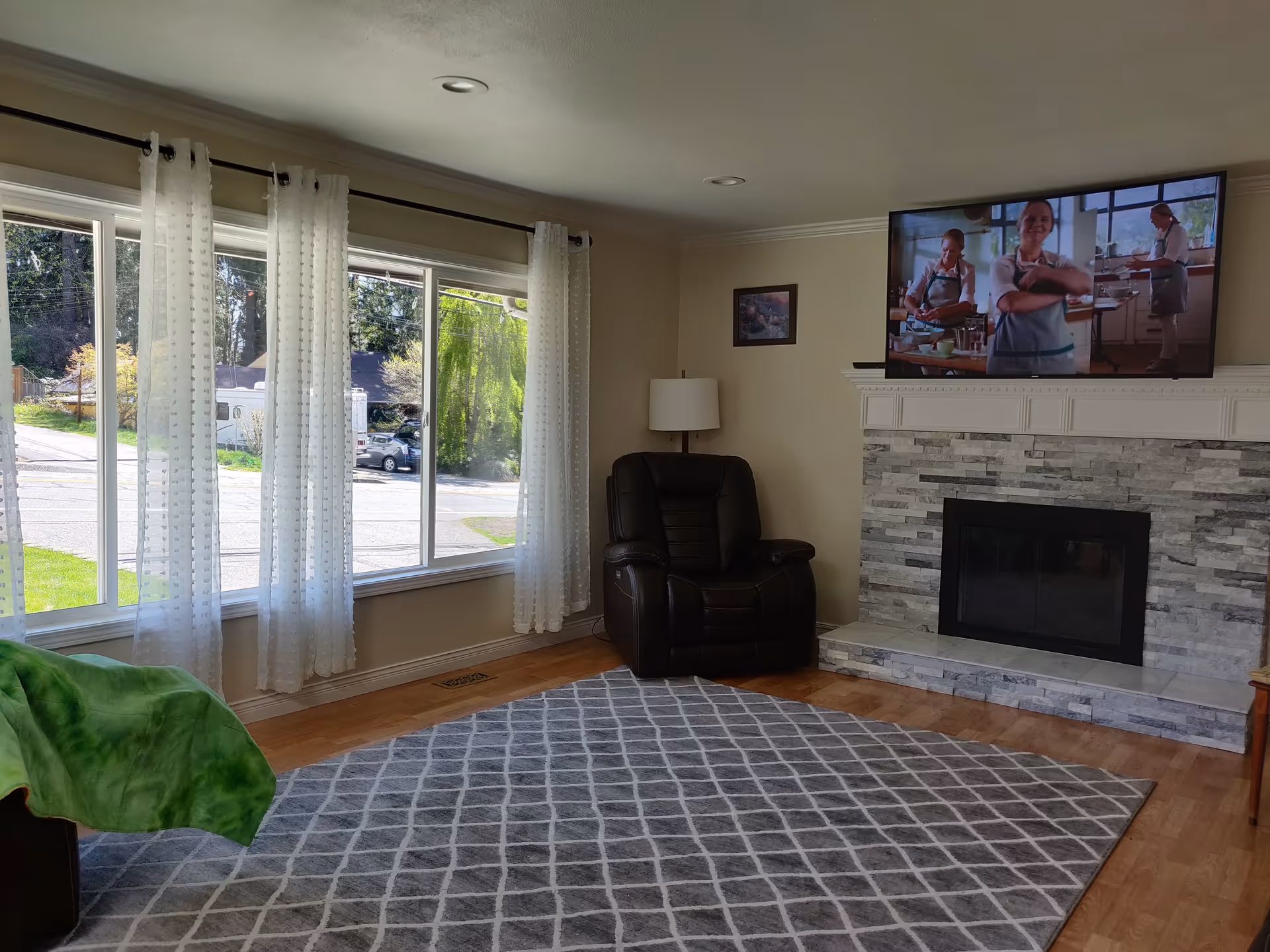 A cozy living room with a large window covered by sheer white curtains, a gray patterned rug on a wooden floor, a black leather recliner chair next to a floor lamp, and a wall-mounted flat-screen TV above a modern stone fireplace.