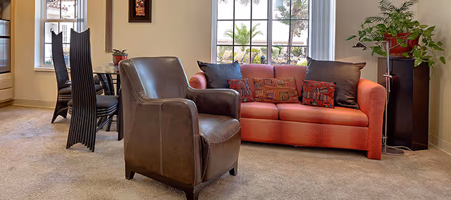 Cozy living area with a red sofa, a brown leather armchair, dining chairs in the background, and a potted plant by a window.