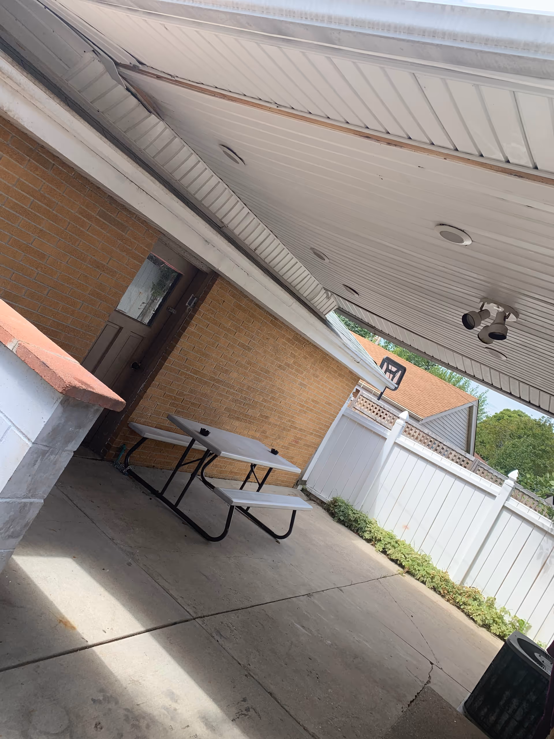 Covered outdoor patio area with a white picnic table and benches on a concrete floor. The area is adjacent to a brick wall with a brown door and a white fence with some greenery along the base. There is a basketball hoop attached to the fence and ceiling lights installed on the white overhead covering.