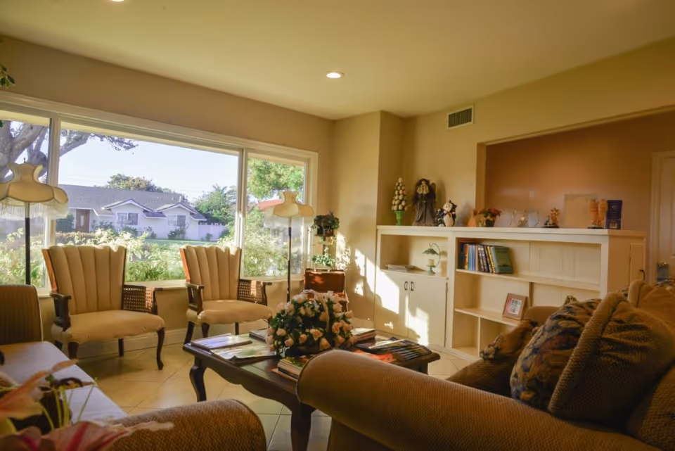 Sunlit living room with upholstered chairs and sofa around a coffee table, built-in shelving, lamps, and a large window overlooking neighboring houses.