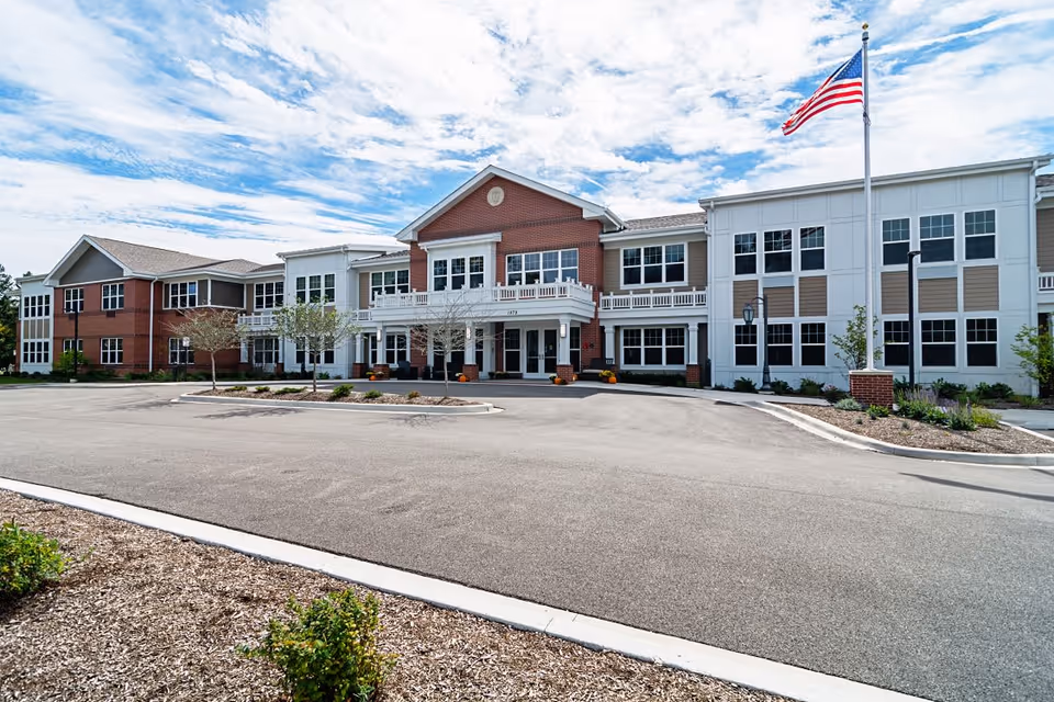 Front exterior view of a large two-story senior living facility building with a combination of red brick and white siding. There is a circular driveway in front, small trees, and landscaping. An American flag is flying on a flagpole near the entrance under a partly cloudy sky.