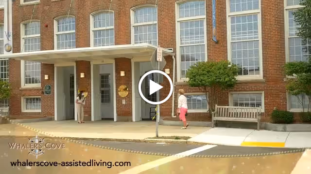 Exterior view of a brick building with large windows and a covered entrance. Two people are standing near the entrance, one on the sidewalk and the other near the door. There is a bench and some greenery near the building. The image includes the text 'Whaler's Cove' and the website 'whalerscove-assistedliving.com' at the bottom left corner.