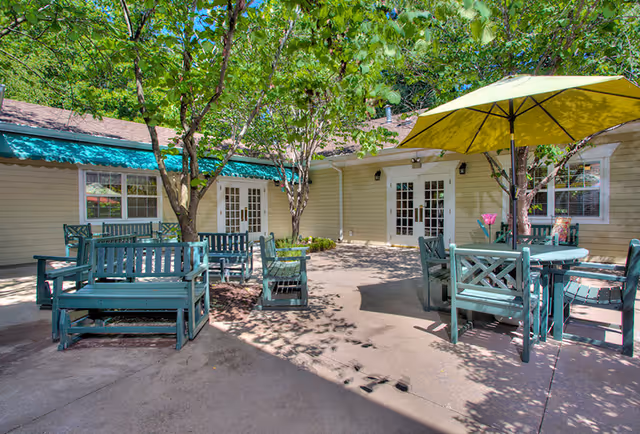 Outdoor patio area at Morningside of Raleigh with green wooden benches and tables, some shaded by trees and a large yellow umbrella. The patio is surrounded by a beige building with multiple white-framed glass doors and windows.