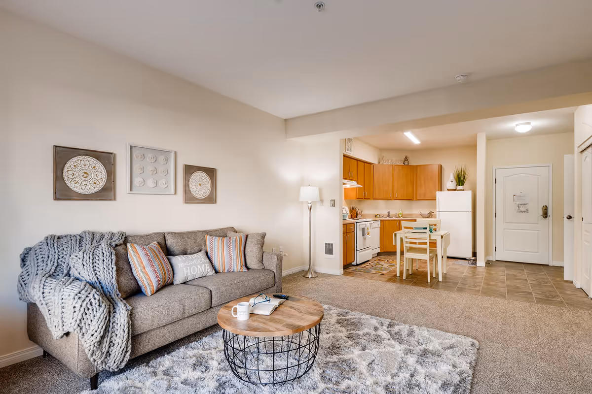 A cozy living room and kitchen area in an apartment at Vancouver Pointe. The living room features a gray sofa with colorful striped pillows and a chunky knit throw blanket, a round wooden coffee table with a book, glasses, and a mug on it, and a fluffy gray rug. The kitchen has wooden cabinets, a white refrigerator, a stove, and a small dining table with two chairs. The walls are decorated with three framed art pieces above the sofa, and a floor lamp stands next to the sofa. The entrance door is visible in the background.