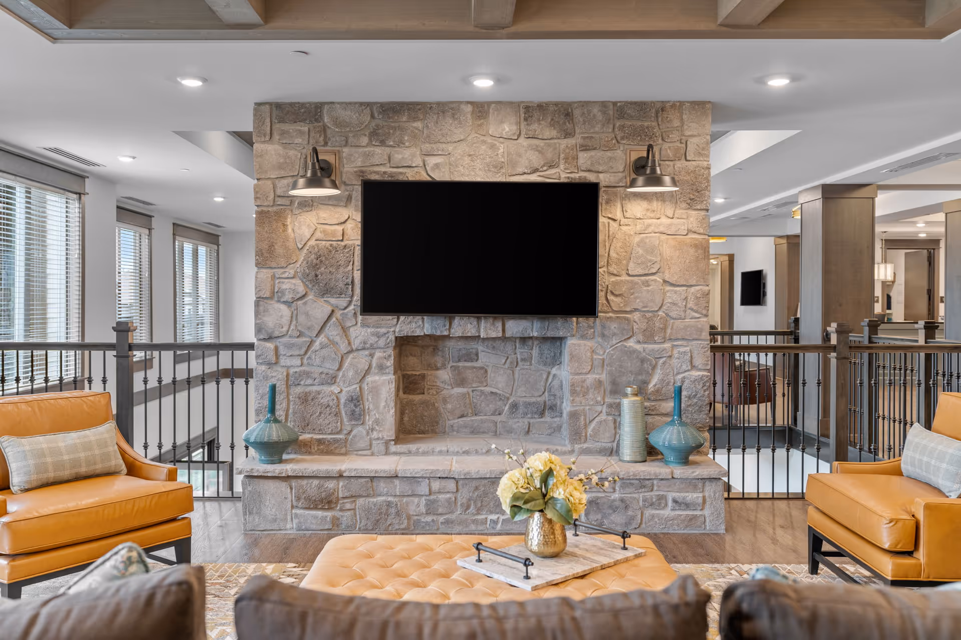 Communal living area with a stone fireplace and mounted flat-screen TV, leather armchairs, an ottoman, and decorative vases.