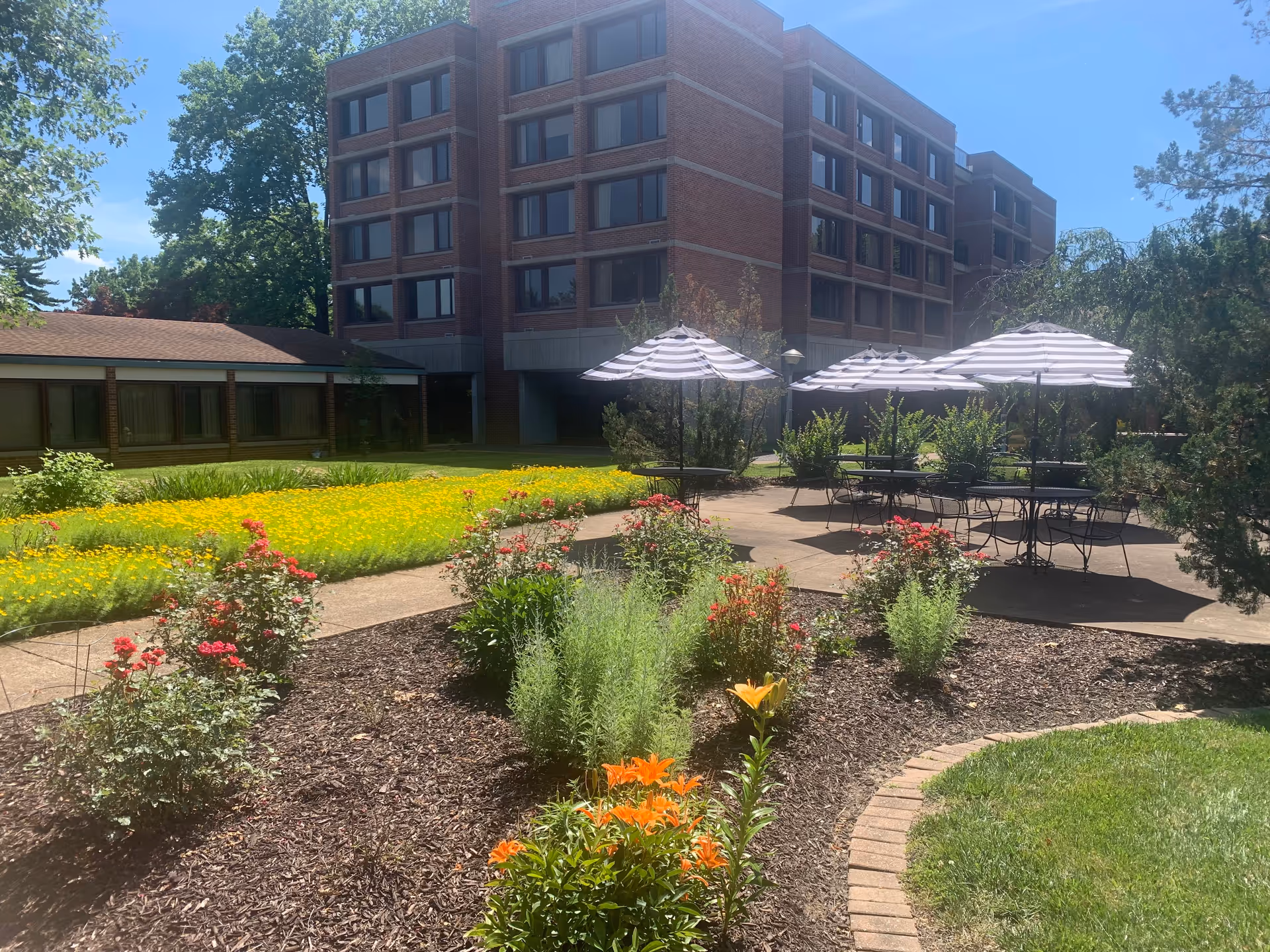Outdoor garden area at Benedictine Living Community-At The Shrine featuring a flower bed with orange and red flowers, a paved patio with black metal tables and chairs under black and white striped umbrellas, and a multi-story brick building in the background under a clear blue sky.
