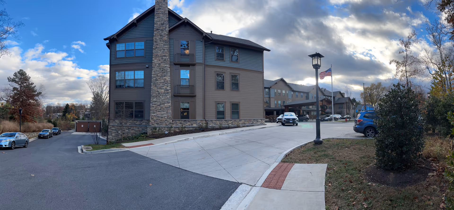 Exterior view of a senior living facility building with a stone chimney, multiple windows, and a driveway leading to the entrance. Several cars are parked along the driveway and in the parking area. An American flag is flying on a flagpole near a streetlamp. Trees and shrubs surround the area under a partly cloudy sky.