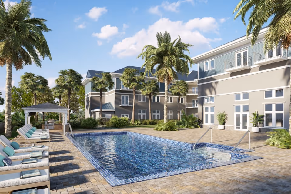 Outdoor swimming pool area at a senior living facility with lounge chairs lined up on the left side, palm trees surrounding the pool, and a covered seating area in the background. The building has multiple floors with balconies and large windows under a partly cloudy blue sky.