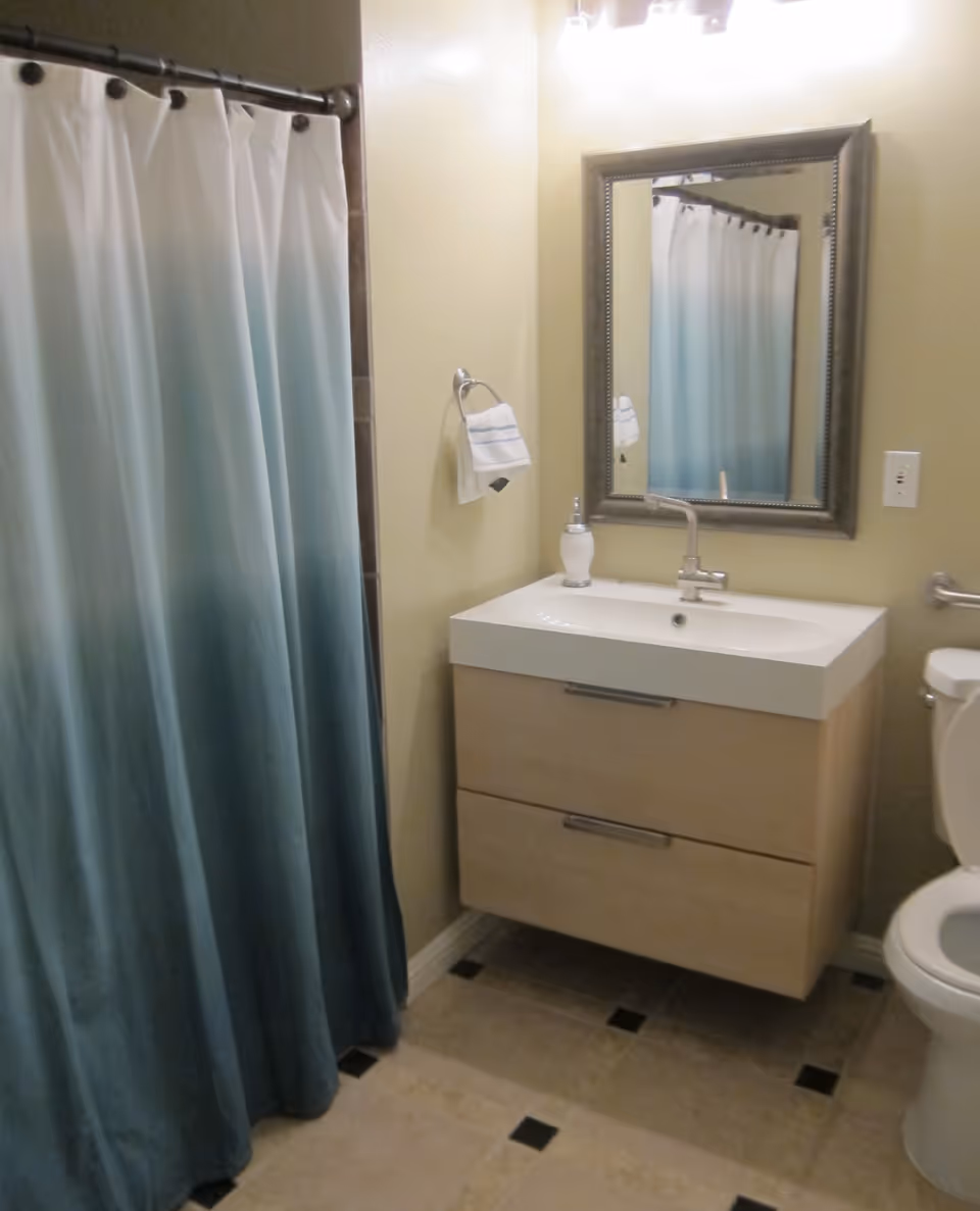 A bathroom with a blue and white gradient shower curtain, a beige floating vanity with a white sink, a silver-framed mirror above the sink, a soap dispenser on the vanity, a towel hanging on a ring, and a toilet partially visible on the right.