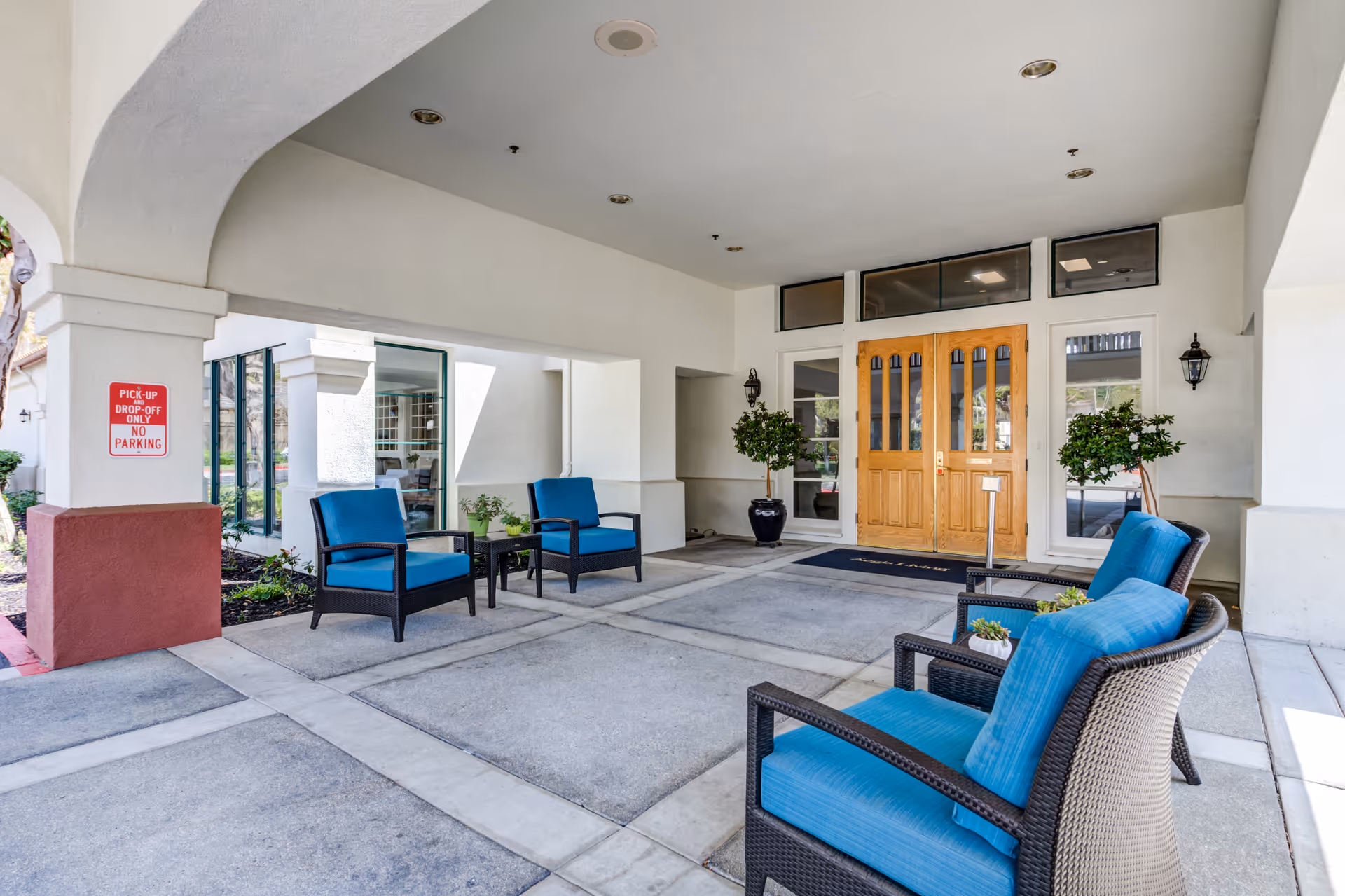 Covered entrance area of a facility with four blue cushioned chairs arranged around small tables, two potted plants near wooden double doors, and a sign indicating pick-up and drop-off only with no parking.