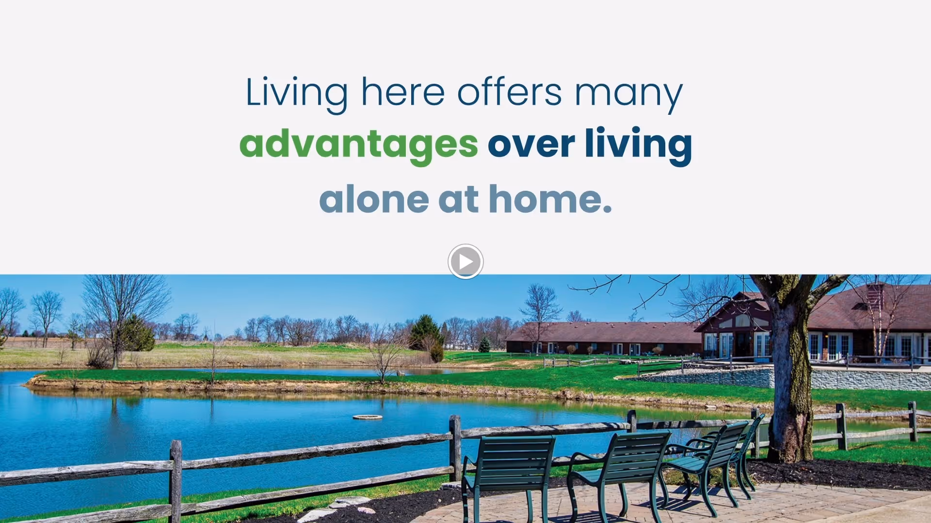 Outdoor view of a retirement community with a pond, wooden fence, green lawn, trees, and benches along a paved walkway. Buildings with brown roofs are visible in the background under a clear blue sky.