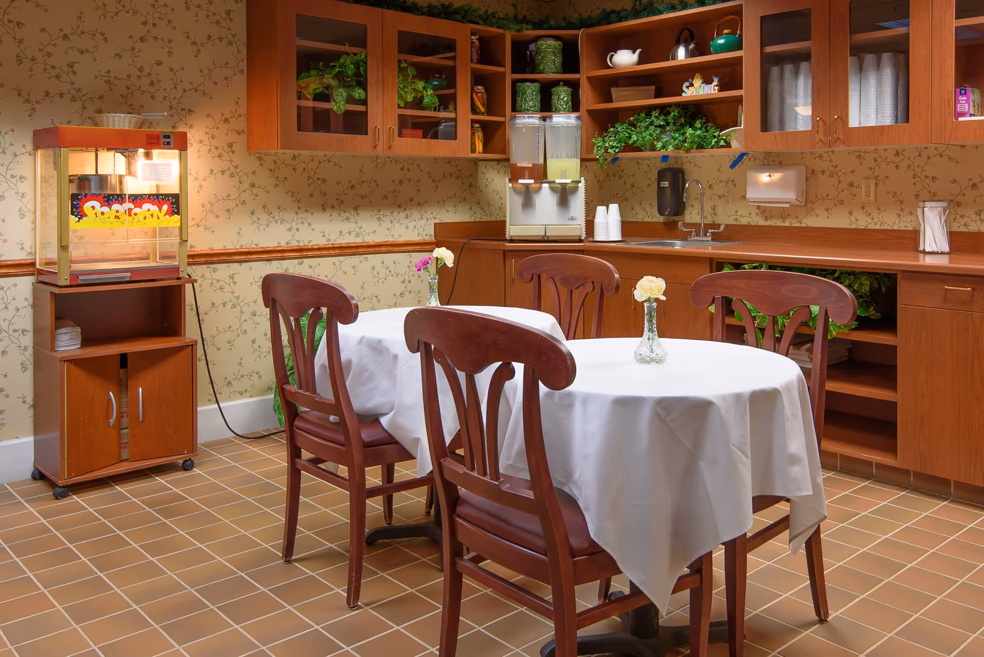 A small dining area with a round table covered with a white tablecloth and four wooden chairs. Each chair has a small vase with a flower on the table. In the background, there is a wooden cabinet with glass doors, a countertop with a sink, two beverage dispensers, and some plants and decorative items on the shelves. To the left, there is a popcorn machine on a wooden stand. The walls have floral wallpaper and the floor is tiled.