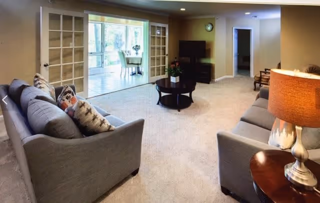 A spacious living room with two gray sofas facing each other, a round coffee table in the center with a plant, a side table with a lamp, and a TV on a stand against the wall. The room has beige walls and carpeted floor. In the background, there is a sunroom or enclosed porch with large windows and a dining table with chairs.
