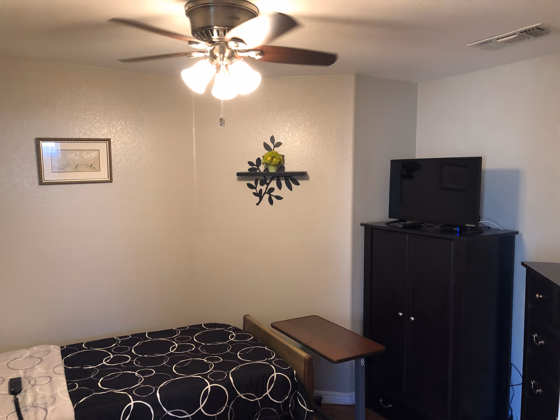 Small furnished bedroom with a single bed, ceiling fan, wall decor, a TV on a dark cabinet, and an overbed table.