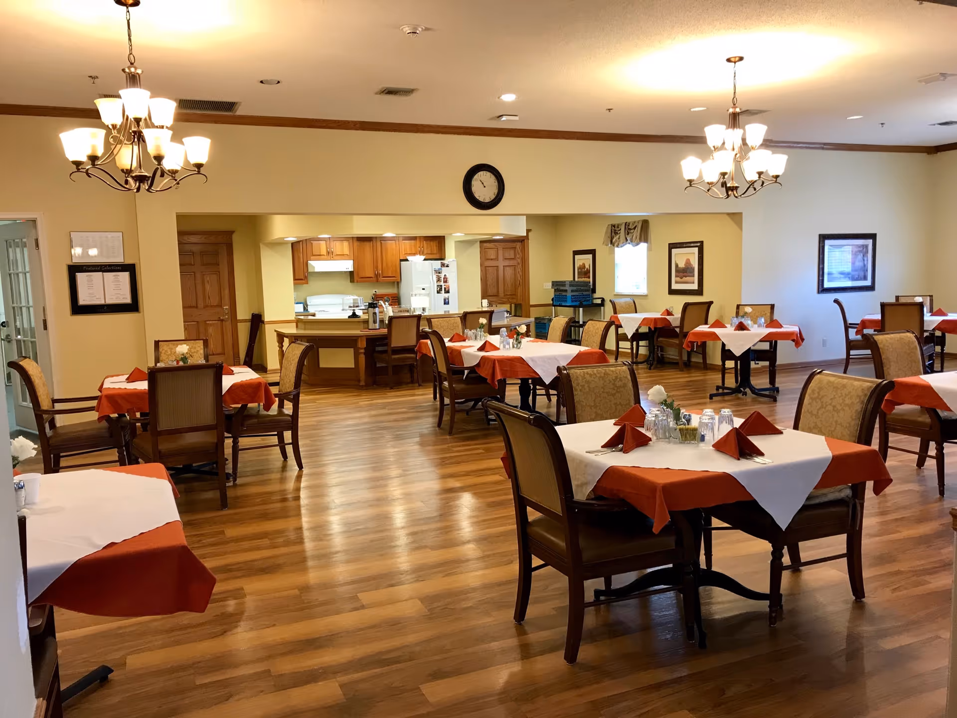 Spacious dining room with multiple tables set with red and white tablecloths and chairs, and a kitchen area visible in the background.