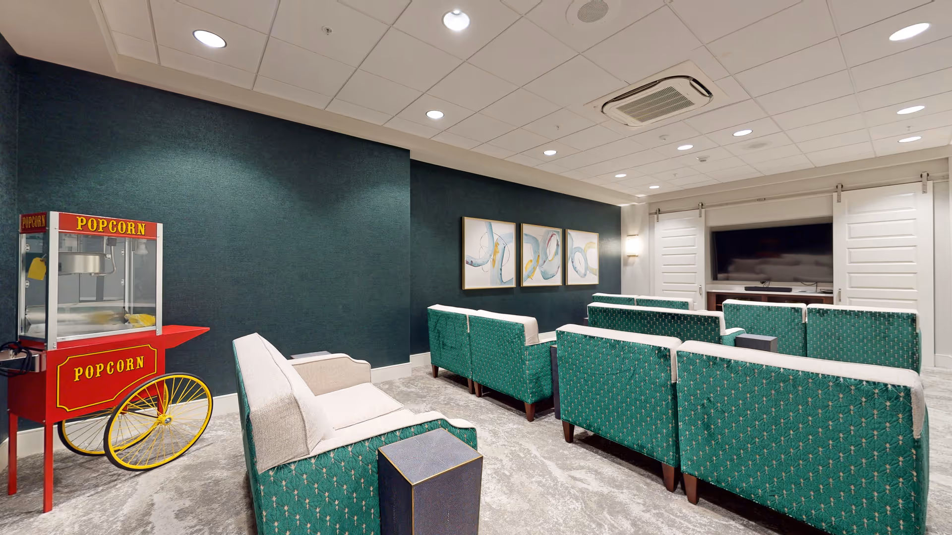 A cozy media room with multiple green and beige upholstered chairs arranged in rows facing a large flat-screen TV mounted on a dark green accent wall. There is a red and yellow popcorn machine on wheels positioned to the left side of the room. The ceiling has recessed lighting and a ceiling-mounted air conditioning unit.