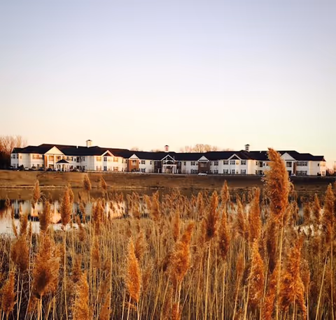 Wide exterior view of a large, two-story assisted living facility building with white walls and dark roof, seen from across a field with tall brown grasses and a small body of water in the foreground during sunset.