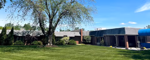 Exterior view of a single-story brick building with a large green lawn and a tree in front under a blue sky with some clouds.