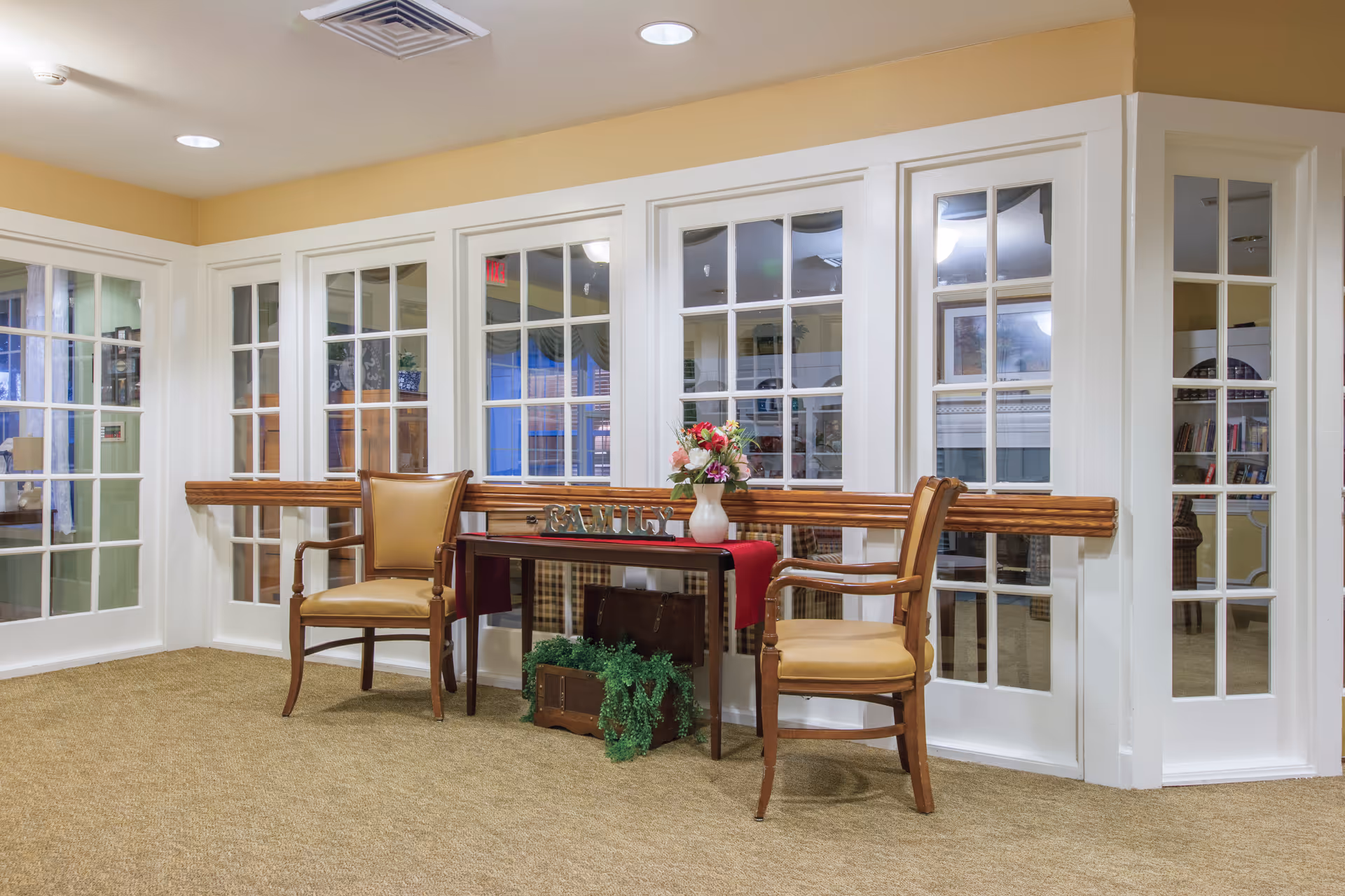 A cozy interior corner with two wooden chairs featuring tan cushions placed on either side of a small table. The table is decorated with a red runner, a vase of flowers, and a decorative 'FAMILY' sign. Behind the table and chairs are large white-framed glass doors and windows, allowing a view into an adjacent room with bookshelves and additional seating. The walls are painted a warm beige color, and the floor is carpeted in a neutral tone.