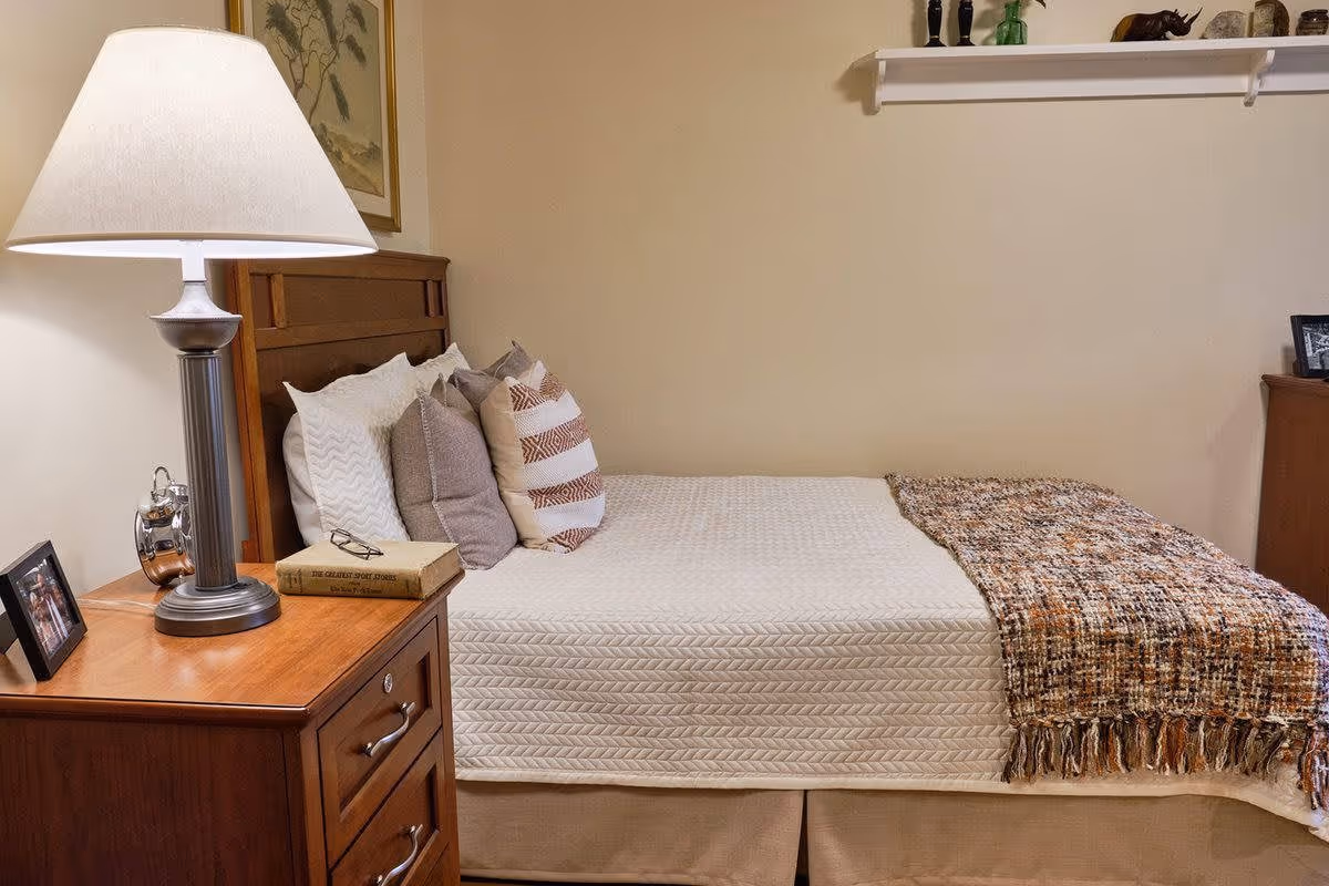 A neatly made bed with a white quilted bedspread and several decorative pillows in neutral tones. A brown and orange patterned throw blanket is draped across the foot of the bed. Next to the bed is a wooden nightstand with a silver lamp, a book, a pair of glasses, and a framed photo. The wall behind the bed is beige with a framed picture and a white shelf holding small decorative items.